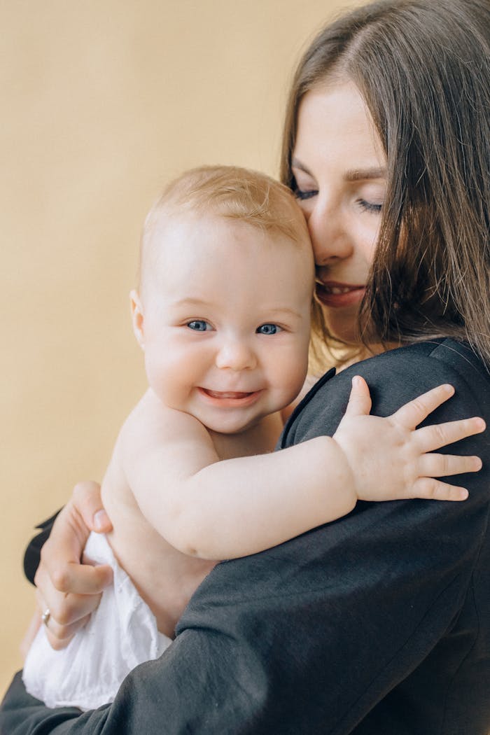 services-02 A joyful mother lovingly holds and hugs her smiling baby, both enjoying a tender moment against a soft yellow backdrop.